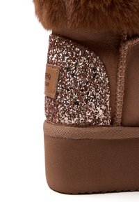 Botín marrón hasta el tobillo con un acolchado de piel de pelo suave y un detalle brillante en oro rosa en el tacón. Textura de ante suave con suela de goma.