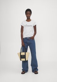 Witte t-shirt met "Armani Exchange" logo-print, blauwe wijde jeans en een strooien tas met zwarte accenten. Model staat tegen een effen achtergrond.