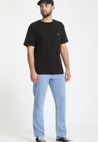 Czarna bawełniana koszulka z krótkimi rękawami, z małym logo. Noszona z jasnoniebieskimi dżinsami i czarnymi trampkami. Gładkie tło.