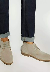 Beige suede desert boots with laces worn with dark rolled-up denim jeans on a neutral background.