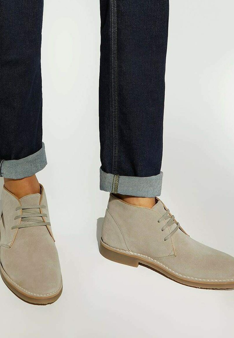 Beige suede desert boots with laces worn with dark rolled-up denim jeans on a neutral background.