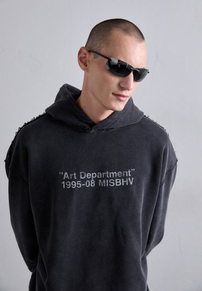 Mann mit kurzen Haaren, der dunkle Sonnenbrillen und einen schwarzen Hoodie mit dem Text "Art Department 1995-08 MISBHV" trägt, vor einem schlichten Hintergrund.