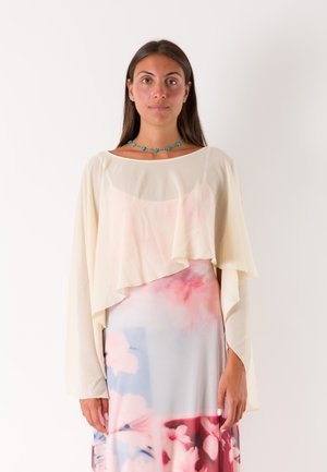 Donna con capelli lunghi e scuri indossa una maglia crema trasparente sopra un vestito floreale rosa e blu e una collana di perle turchese, in piedi su sfondo bianco.