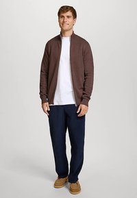 Braune Reißverschlussjacke mit Chevron-Struktur, getragen über einem weißen T-Shirt. Kombiniert mit dunkelblauen Hosen und hellbraunen Slippern.