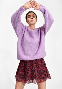 Lila oversized gebreid sweater met ribbelkraag en -mouwen, gecombineerd met een knielange rok met bloemendruk in rode, roze en blauwe tinten.