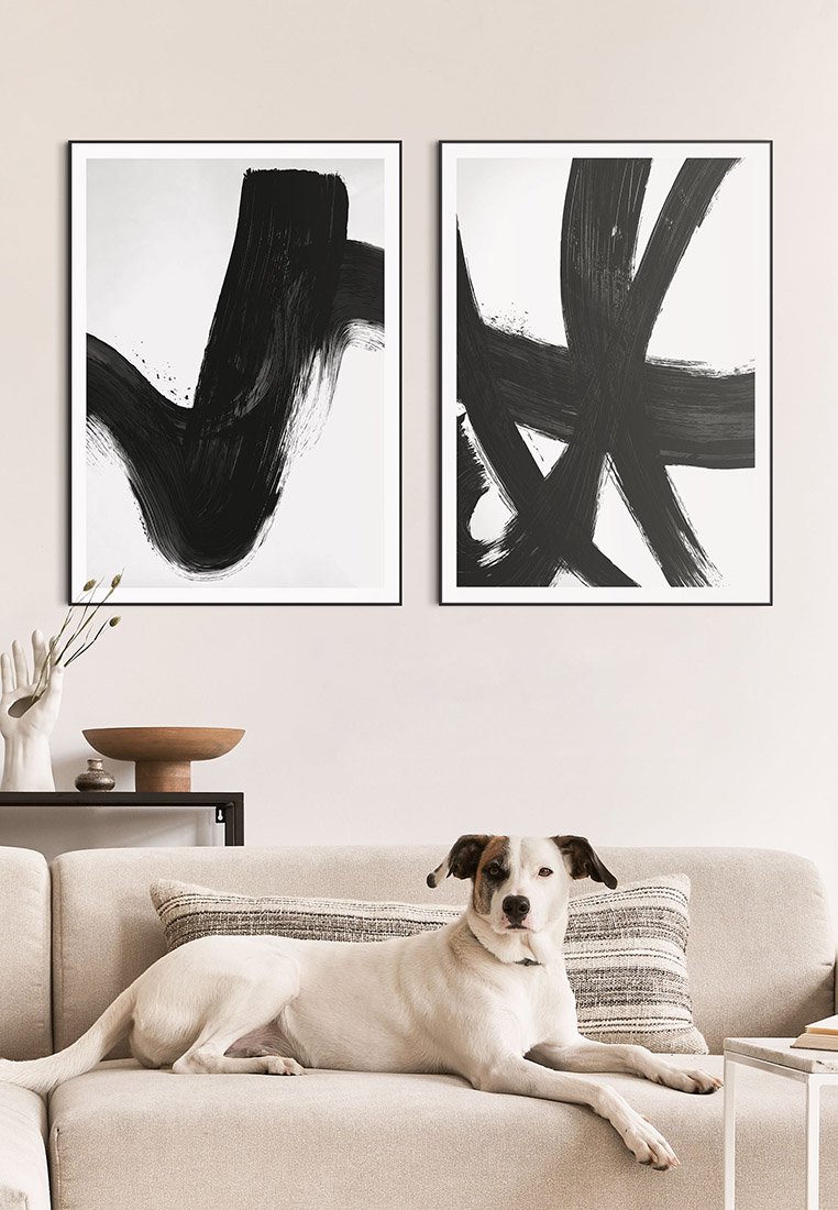 Zwei schwarz-weiße abstrakte Gemälde in Rahmen über einem beigen Sofa mit strukturierten Kissen und einem Hund, der darauf liegt. Einfache Dekorationselemente sind sichtbar.