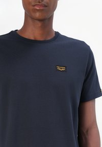 Navy blue cotton t-shirt with a round neckline and short sleeves. Features a small black and gold "PME Legend" logo patch on the chest.
