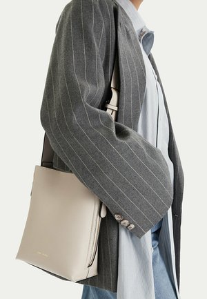 Persona que lleva una chaqueta de raya diplomática gris y una camisa azul claro, y lleva un bolso de cuero beige con cremallera en el lado izquierdo.
