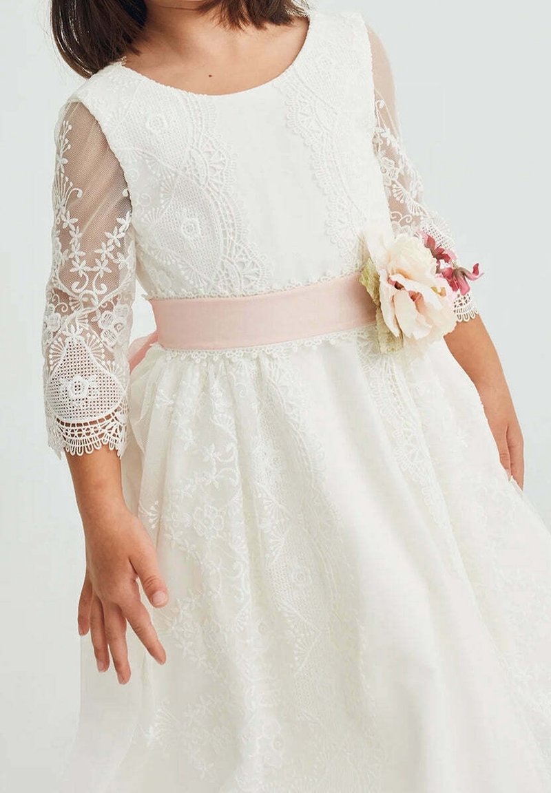 Fille portant une robe en dentelle blanche avec manches transparentes brodées et ceinture rose ornée de fleurs en tissu, debout devant un fond uni.