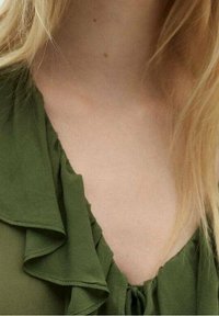 Blondes Haar über die Schultern, trägt eine grüne Bluse mit Rüschenkragen und V-Ausschnitt.