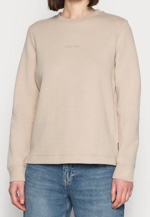 Beiger Sweatshirt mit langen Ärmeln und dezentem Calvin Klein-Logo auf der Brust, kombiniert mit mittelblau gewaschenen Jeans.