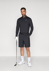 Polo noir à manches longues avec col et logo Nike, associé à un short noir. Accessoires incluent un club de golf et un gant de golf blanc.