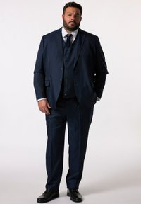 Traje de tres piezas en azul marino con solapa muesca, chaleco de seis botones y pantalones rectos, hecho de una tela texturizada. Zapatos de vestir negros.