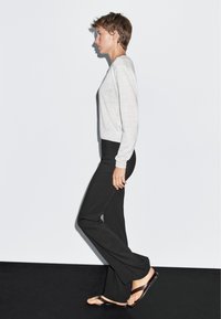 Hellgrauer Pullover mit langen Ärmeln, schwarze ausgestellte Hose und braune Flip-Flops. Das Outfit besticht durch eine lockere Passform und weiche Texturen.