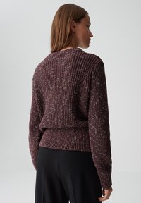 Frau mit glattem braunem Haar, die einen kastanienbraunen melierten Strickpullover und schwarze Hosen trägt, seitlich vor einem schlichten grauen Hintergrund.
