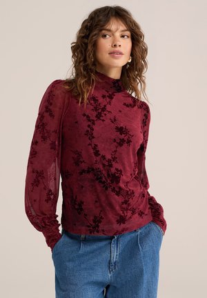 Femme aux cheveux bruns ondulés portant un haut à manches longues bordeaux transparent à motif floral et un jean bleu, posant mains dans les poches devant un fond beige.