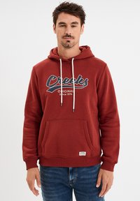 Sudadera burdeos con cordones, bolsillo frontal tipo canguro y un gráfico que dice "Creeks Original Desde 1974" impreso en el pecho. Textura de tejido suave.