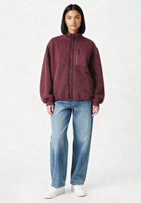 Giacca in pile bordeaux con collo alto, zip integrale e tasche laterali; presenta una finitura testurizzata e un pannello in tessuto liscio a contrasto.