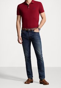 Czerwona koszulka polo z małym logo, niebieskie dżinsy, brązowe skórzane buty i brązowy pasek. Model stoi z rękami w kieszeniach.