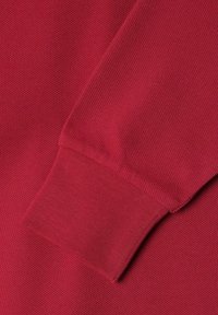 Roter strukturierter Stoff mit einem gerippten Bündchen und einer diagonalen Naht. Die Oberfläche zeigt ein dezentes gewebtes Muster.