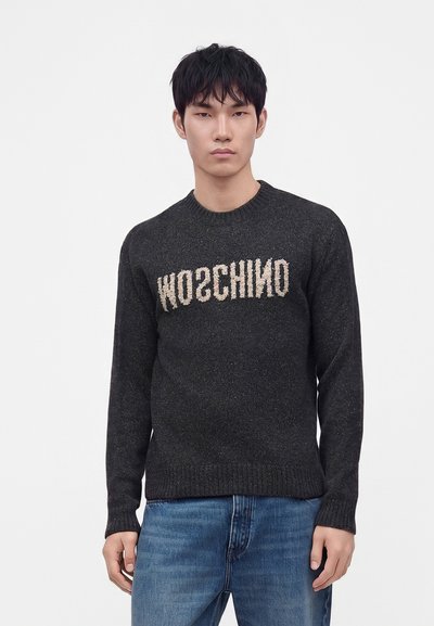 Jeune homme aux cheveux courts et foncés, portant un pull noir avec un texte beige inversé "MOSCHINO" et un jean bleu, debout devant un fond blanc.
