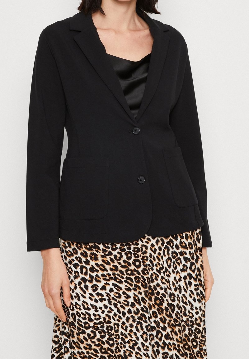 Femme portant un blazer noir boutonné sur un haut noir, assorti d'une jupe à imprimé léopard, debout devant un fond uni.