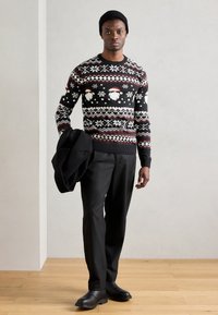 Suéter preto de malha com padrões festivos de Pai Natal e flocos de neve, combinado com calças de fato pretas e um gorro preto, segurando um casaco.