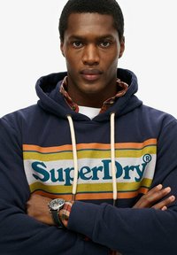 Granatowa bluza z kapturem SuperDry z dużym logo, wielokolorowymi paskami, ściągaczem oraz ściągaczami na rękawach. Noszona na koszuli w kratę. Srebrny zegarek na nadgarstku.