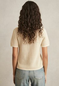 Maglione a maglia con maniche corte, colore crema, texture a coste e design aderente. Abbinato a jeans blu chiaro, visuale di schiena.