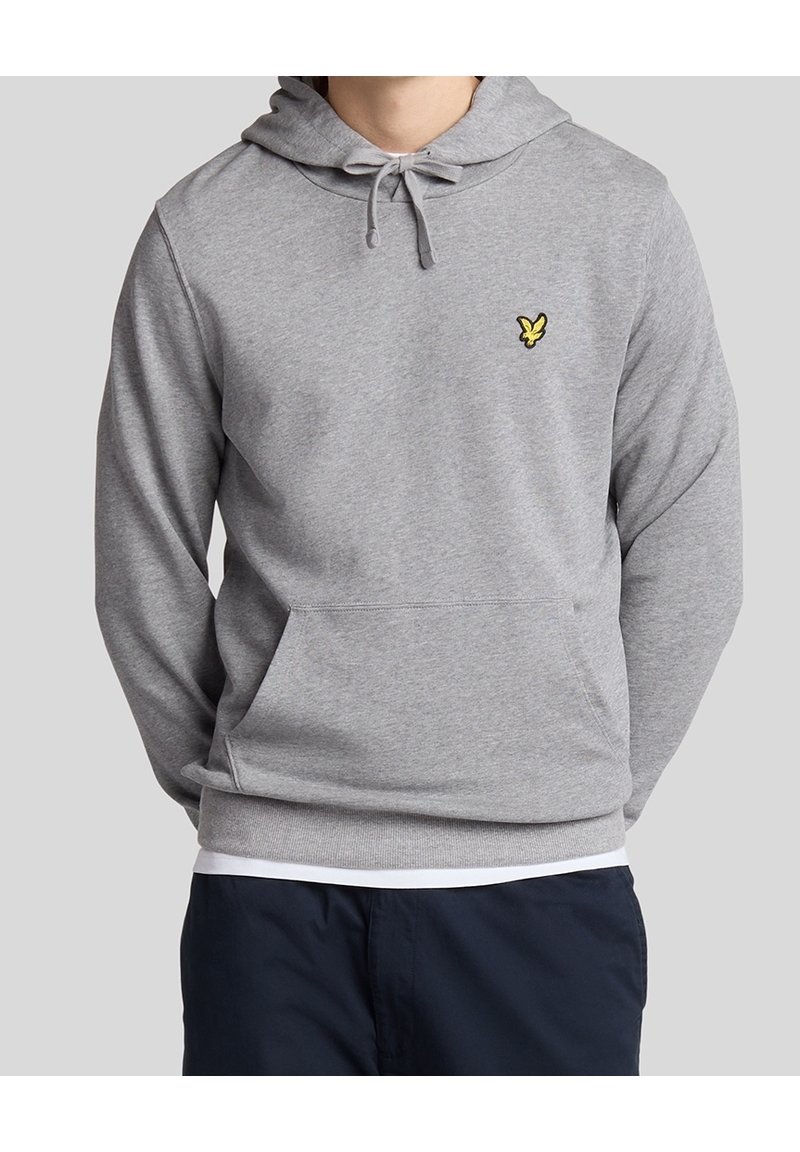 Homme portant un sweat à capuche gris clair avec une poche avant et un petit logo d'oiseau jaune sur la poitrine, associé à un pantalon foncé.