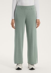 Světlé zelené, žebrované kalhoty s širokými nohavicemi, vyrobené z měkkého materiálu a s plochým páskem. Spojené s bílými sportovními botami.