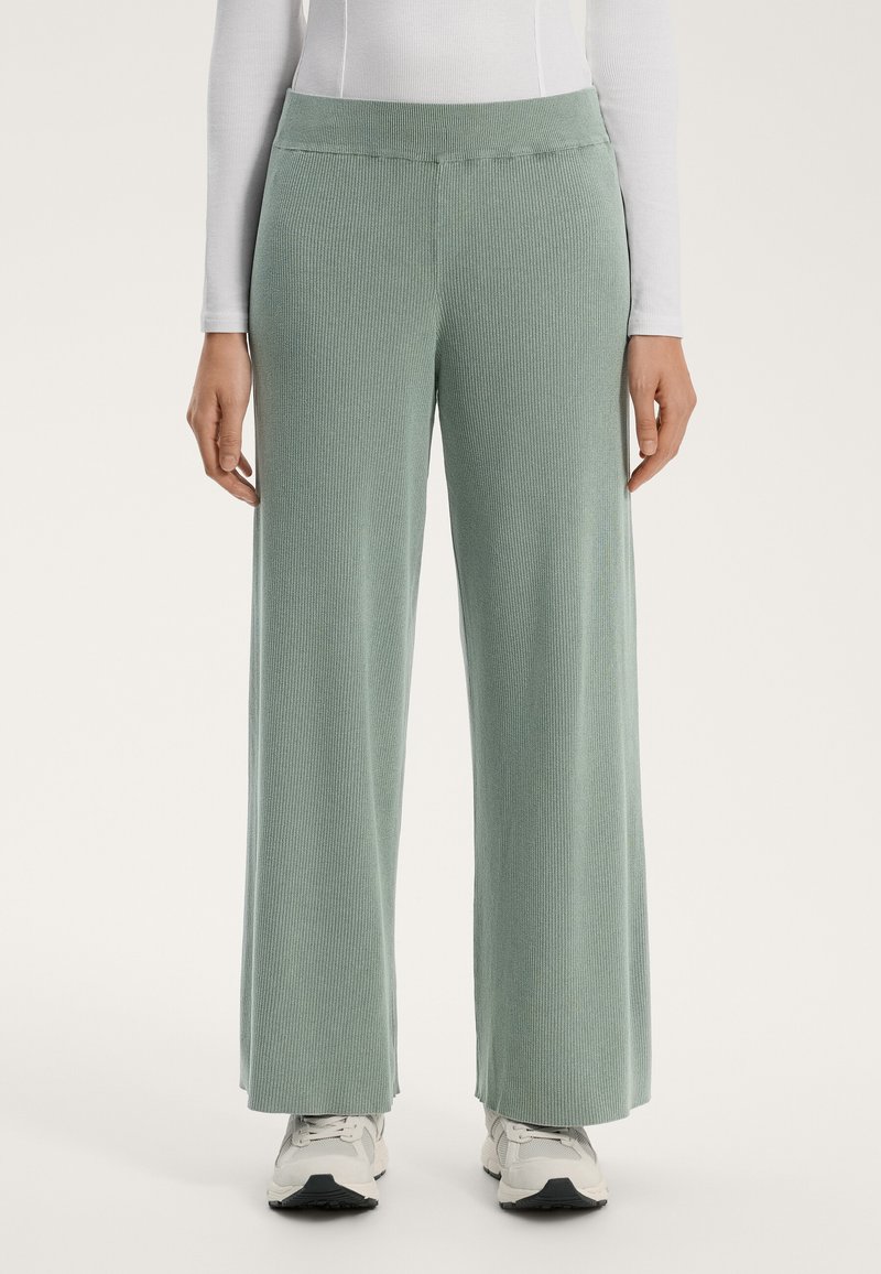 Světlé zelené, žebrované kalhoty s širokými nohavicemi, vyrobené z měkkého materiálu a s plochým páskem. Spojené s bílými sportovními botami.