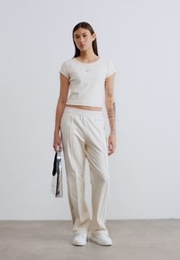 Λευκό κοντοκομμένο t-shirt με μικρό λογότυπο, σε συνδυασμό με λευκή φόρμα σε χαλαρή γραμμή. Το μοντέλο κρατά μια ασημένια τσάντα και φοράει λευκά παπούτσια.