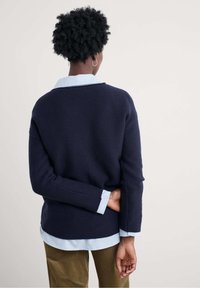 Maglione navy con vestibilità rilassata, polsini a costine e una camicia a strisce azzurro chiaro sovrapposta. Texture morbida, design minimal.