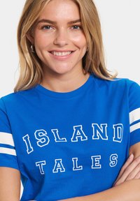 T-shirt en coton bleu avec des lettres blanches en bloc portant l'inscription "ISLAND TALES" et des rayures blanches sur les manches. Texture lisse, coupe décontractée.