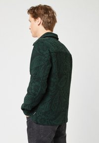 Giovane uomo con capelli corti castani che indossa una giacca a motivo verde scuro e jeans neri, rivolto a sinistra con le mani nelle tasche.