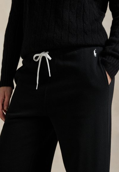 Person iført sort kabelstrikket sweater og sorte joggingbukser med hvid snor og lille logo på hoften, hånd i lommen.