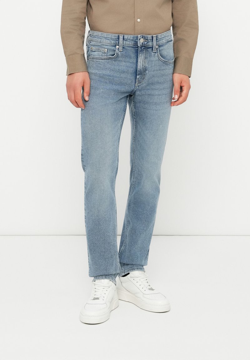 Jeans din denim de culoare albastru deschis, cu o croială dreaptă, design cu cinci buzunare, accesorii metalice și detalii de cusătură subtile, purtați cu adidași albi.