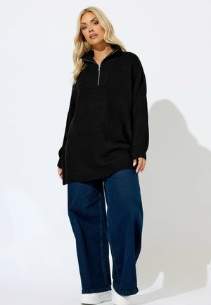 Schwarzer gestrickter Pullover mit einem halben Reißverschluss, langen Ärmeln und einem lockeren Schnitt, kombiniert mit weiten, dunkelblauen Jeans und weißen Sneakern.