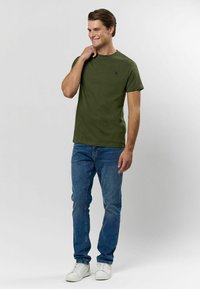 Olivově zelené tričko s kulatým výstřihem vyrobené z bavlny, v kombinaci s modrými džínami a bílými teniskami, stojící na světlém pozadí.