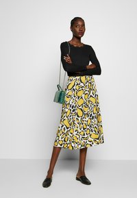 Maglietta nera a maniche lunghe, gonna midi leopardata giallo e nera, scarpe slip-on nere e una piccola borsa a tracolla verde. Texture liscia.