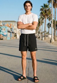 Mladi moški stoji z rokami prekrižanimi na sončni sprehajalni poti ob plaži, obdani z datlji in pisanimi zgradbami.