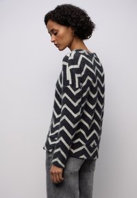 Svetr s cikcak vzorem v tmavě šedé a krémové barvě, vyrobený z měkkého, texturovaného materiálu. Volný střih s bočními rozparky.