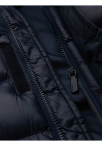 Acercamiento a la cremallera de una chaqueta acolchada de color azul marino oscuro y a la tira de Velcro negra adyacente en la tela acolchada.