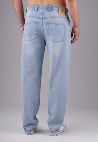 Mann in hellblauen Jeans und weißen Converse-Sneakern, stehend mit dem Rücken zur Kamera vor einem schlichten Hintergrund.