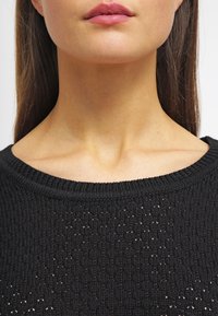 Kvinnas nedre ansikte, nacke och övre bröst synliga, klädd i en svart stickad tröja med rund halsringning och rakt brunt hår.