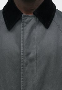 Grå jakke med en ribbet krave, der har en glat tekstur og synlige syninger langs frontknaplisten. Minimalistisk design.