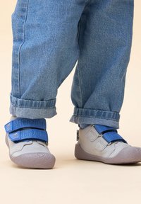 Grijze schoenen met blauwe klittenbandsluitingen, met een gestructureerde rubberen zool, gedragen met opgerolde spijkerbroek.