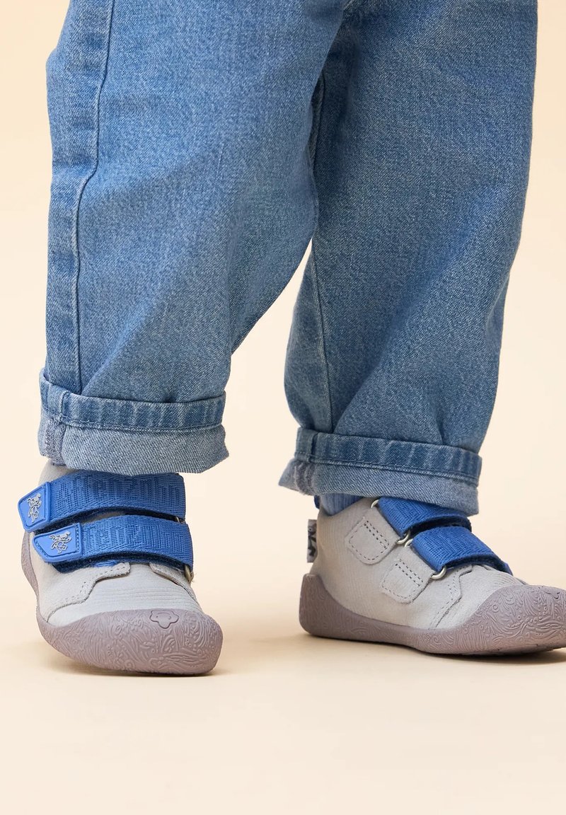 Grijze schoenen met blauwe klittenbandsluitingen, met een gestructureerde rubberen zool, gedragen met opgerolde spijkerbroek.