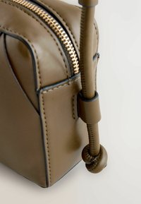 Sac à main en cuir marron avec une surface texturée, une fermeture éclair dorée et une anse nouée en accent. Présente une couture noire autour des bords.
